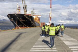 Personas con chaleco de seguridad caminando en un puerto con un barco carguero amarrado